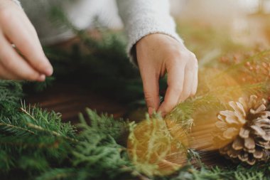 Yeşil köknar dallarını tutan ve arka planda çam kozalakları, böğürtlen ve şenlik ışıklarıyla kırsal ahşap masa üzerinde kırsal Noel çelengi yapan kadınların ellerini kapatın. El yapımı şenlik dekoru