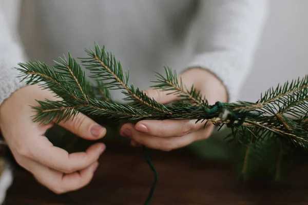 緑のモミの枝を保持し 木製のテーブルの背景に素朴なクリスマスの花輪を作る女性の手の閉じる 手作りのお祝いの装飾 休日の到来 季節の冬のワークショップ — ストック写真