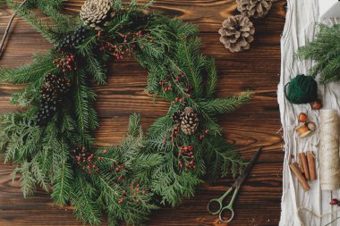 Ahşap masa üzerinde kırsal noel çelengi. Kırmızı böğürtlenli modern Noel çelengi, yeşil dallar, çam kozalakları, makas, kırsal arka planda sicim. Tatil atölyesi