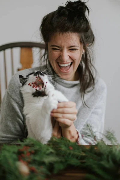 若い幸せな女性が素朴なクリスマスリースを作るのを助けるかわいい猫は 赤い果実と緑の枝を保持します 面白い本格的な家庭の瞬間 ハッピーホリデー 助っ人と笑う女 — ストック写真