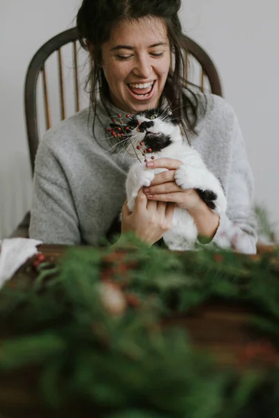 若い幸せな女性が素朴なクリスマスリースを作るのを助けるかわいい猫は 赤い果実と緑の枝を保持します 面白い本格的な家庭の瞬間 ペットや休日 助っ人と笑う女 — ストック写真