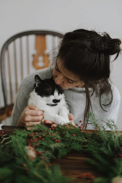 若い幸せな女性が素朴なクリスマスリースを作るのを助けるかわいい猫は 赤い果実と緑の枝を保持します 面白い本格的な家庭の瞬間 ペットや休日 ハッピー ホリデー — ストック写真