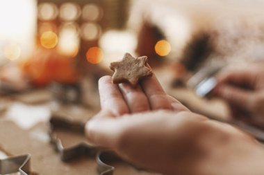 Elinde yıldız zencefilli kurabiye tutan kişi, Noel tatilinin gelişi. Kırsal masadaki yıldız metal kesiciyle zencefilli ekmek hamuru, baharatlar, şenlikli süslemeler, ışıklar.