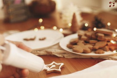 Zencefilli kurabiyeyi ahşap masa üzerindeki kremayla süslemek. Noel tatili geleneği ve gelişimi. Elleri, fırında pişmiş noel yıldızı kurabiyelerini şekerli şekerlemeyle süslüyordu. Aile zamanı.