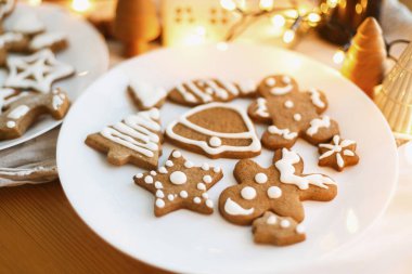 Beyaz tabakta kremalı zencefilli kurabiyeler ahşap masada festival ışıklarıyla. Süslü Noel ağacı, yıldız ve geyik kurabiyeleri şekerli. Noel tatili geleneği ve gelişimi