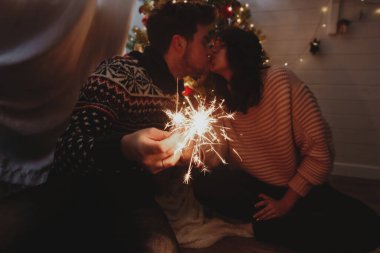 Karanlık festival odasında ışıkları olan bir Noel ağacının arka planında yanan kıvılcımlar tutan şık mutlu çift. Genç bir aile yeni yılı havai fişeklerle kutluyor. Mutlu yıllar.!