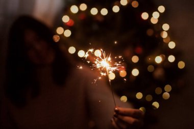 Karanlık festival odasında ışıkları olan bir Noel ağacının arka planında yanan maytap tutan şık, mutlu bir kadın. Genç bayan yeni yılı havai fişeklerle kutluyor. Mutlu yıllar.!