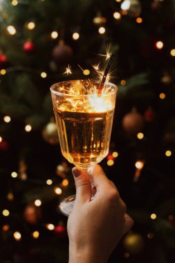 Mutlu yıllar! Karanlık festival odasında ışıkları olan bir Noel ağacının arka planında elinde şampanya bardağıyla havai fişek yakmak. Yeni yılı kutlamak, şık estetik imaj.