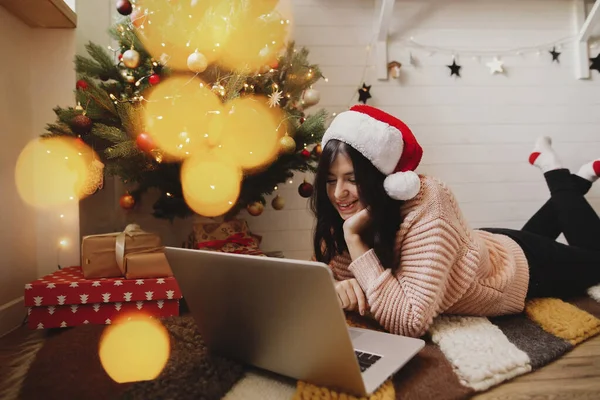 Boldog Fiatal Télapó Kalapban Fekszik Laptoppal Karácsonyfa Alatt Ajándékokkal Fényekkel — Stock Fotó