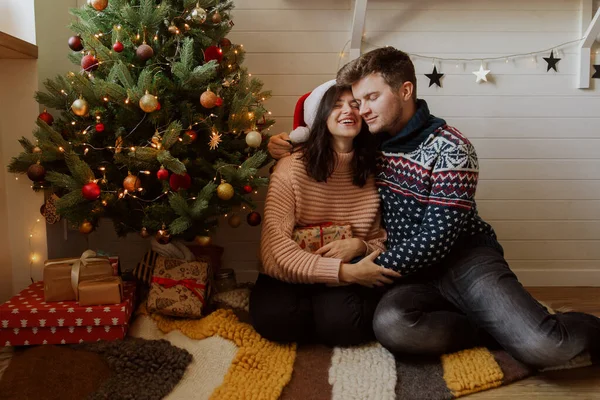 お祝いの装飾された部屋のライトとクリスマスツリーの下でクリスマスプレゼントを保持スタイリッシュな幸せなカップル プレゼントや抱擁 魔法の瞬間を交換する若い家族 メリークリスマス — ストック写真