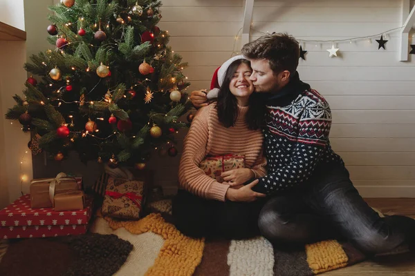 メリークリスマス お祝いの装飾された部屋のライトとクリスマスツリーの下でクリスマスプレゼントを保持スタイリッシュな幸せなカップル プレゼントやキス 魔法の瞬間を交換する若い家族 — ストック写真