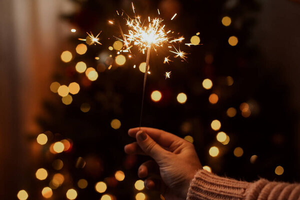 Burning sparkler in hand on background of christmas tree with lights in dark festive room. Young female celebrating new year with firework. Happy New Year!