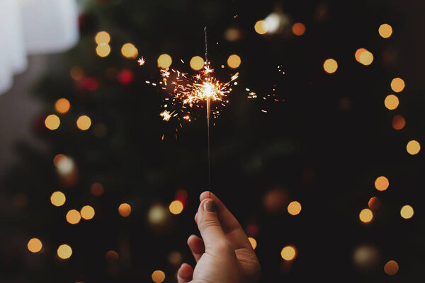 Happy New Year! Burning sparkler in female hand on background of christmas tree with lights in dark festive room. Celebrating new year with firework, stylish aesthetic image.