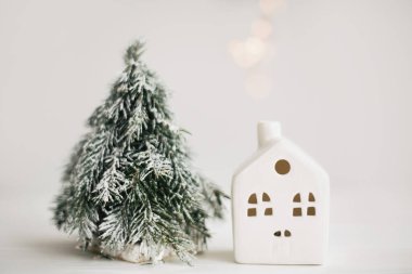 Sıcak kışlık ev, noel sahnesi, ışıklı minyatür ev ve karlı ağaç. Noel ışıkları, küçük seramik ev ve beyaz arka planda köknar ağacı. Mutlu Noeller! Şenlikli modern dekorasyon