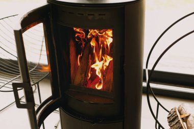 Büyük pencerenin arkasında yanan şöminesi ve metal tezgahında yakacak odunu olan modern siyah şömine. Soğuk mevsimde sıcak ve sakin anlar, kabinde ısıtıcı