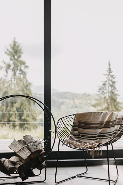 山の景色を望む大きな窓の背景に金属製のスタンドに居心地の良い毛布と薪と現代の黒い椅子 寒い季節にキャビンで居心地の良い暖かく穏やかな瞬間 — ストック写真