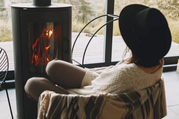 穿着针织毛衣 头戴帽子的时尚女人在山上的现代黑色壁炉前热身 寒冷季节温暖舒适的时刻 嬉皮士女人在舒适的家休息 — 图库照片