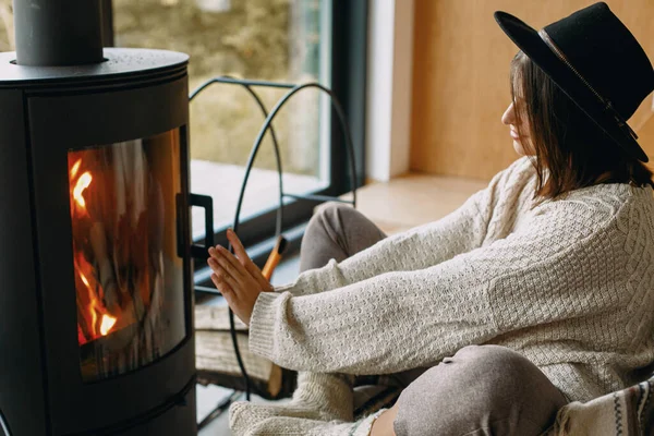 Stylish woman in knitted sweater and hat warming up at modern black fireplace with view on mountains. Cozy warm moments at cold season. Hipster female relaxing in comfortable home