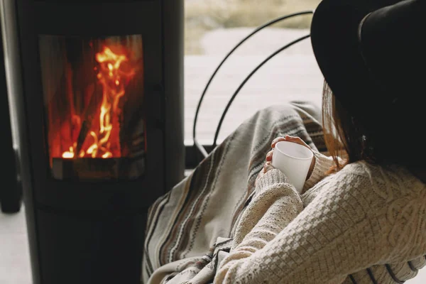 一个时髦的女人 手里拿着一杯热茶 在可以俯瞰群山的现代黑色壁炉边放松一下 寒冷季节温暖舒适的时刻 穿着针织毛衣 头戴帽子的年轻女性 — 图库照片
