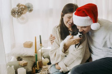 Mutlu, genç bir aile, süslü püslü bir odada oturup sevimli kedileri öpüyor. Mutlu Noeller! Işıklı kedicik ve Noel Baba şapkalı şık çift Noel tatilini evde kutluyor..