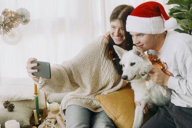 Işıklı köpek yavrusu ve Noel Baba şapkalı şık çift Noel tatilini ailesiyle internetten kutluyor. Video odasında sevimli köpekli mutlu bir aile mi yoksa telefonda dekorlu selfie mi çekiyorlar?