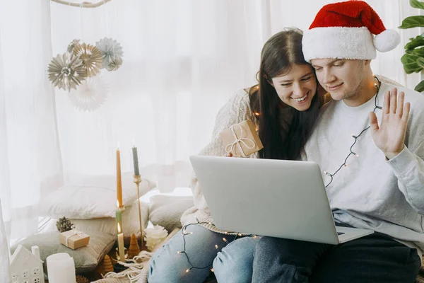 幸せな若いカップルは ノートパソコンでのビデオ通話で家族や友人とプレゼントを交換します ライトとサンタの帽子交換の贈り物でスタイリッシュな家族は お祝いの装飾が施されたブーホールームでオンラインで — ストック写真