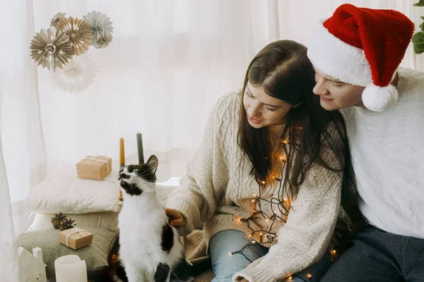 幸せな若い家族が座って お祝いの装飾されたブースでかわいい猫を愛撫 メリークリスマス 光の中で子猫と家でクリスマスの休日を祝うサンタの帽子を持つスタイリッシュなカップル — ストック写真
