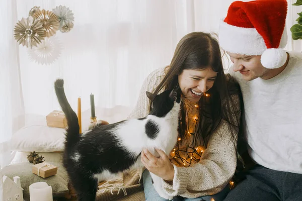 幸せな若い家族が座っているとお祝いの装飾されたブースルームでかわいい猫にキス メリークリスマス 光の中で子猫と家でクリスマスの休日を祝うサンタの帽子を持つスタイリッシュなカップル — ストック写真