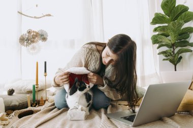 Stylish woman in cozy sweater putting santa hat on cute cat at christmas furoshiki gift and laptop in festive decorated boho room. Young female playing with furry friend. Happy Holidays! clipart