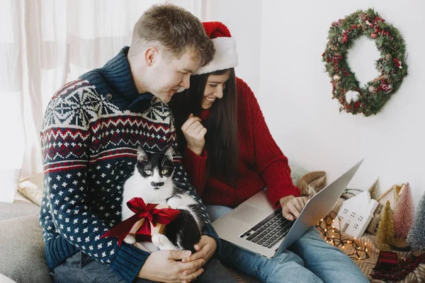 ラップトップを使用してかわいい猫とクリスマスプレゼントと幸せな若い家族とお祝いの部屋で笑顔 ビデオ通話で自宅でクリスマスを祝う セーターやサンタの帽子ショッピングでスタイリッシュなカップルオンライン — ストック写真