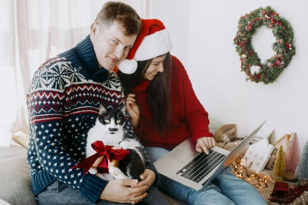 ラップトップを使用してかわいい猫とクリスマスプレゼントと幸せな若い家族とお祝いの部屋で笑顔 ビデオ通話で自宅でクリスマスを祝う セーターやサンタの帽子ショッピングでスタイリッシュなカップルオンライン — ストック写真