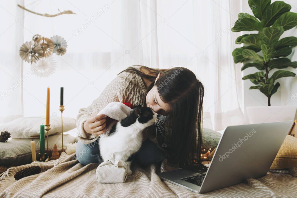 Stylish woman in cozy sweater caressing and kissing cute cat in  santa hat at christmas furoshiki gift and laptop in festive decorated boho room. Celebrating at home, Merry Christmas!