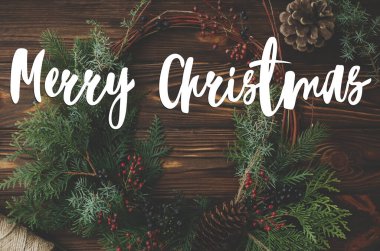 Modern rustik noel çelengi üzerine yazılmış mutlu noel metinleri ahşap masa üzerinde kırmızı böğürtlenler ve çam kozalaklarıyla yatıyordu. Tebrik kartı, modern mevsimler selamları. İyi tatiller.