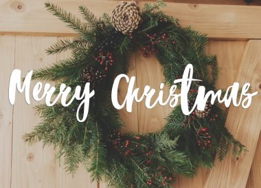 Ahşap kapıda el yazısıyla Noel çelengi ve bayram dekorasyonu üzerine Mutlu Noeller mesajı. Tebrik kartı, modern mevsimler selamları. İyi tatiller.