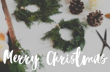 Mutlu Noeller tebrik kartı. Kırsal Noel çelenginde mumlarla, kozalaklarla ve beyaz ahşap süslemelerle el yazısıyla mutlu Noeller mesajı. Mutlu tatiller, mevsimler selamlar