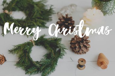 Kırsal Noel çelenginde mumlarla, kozalaklarla ve beyaz ahşap süslemelerle el yazısıyla mutlu Noeller mesajı. Tebrik kartı, modern mevsimler selamları. İyi tatiller.
