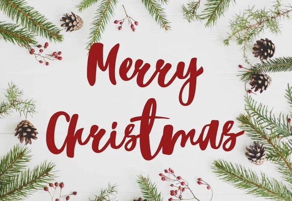 圣诞快乐 圣诞快乐的文字手写在枞树枝上 红色的浆果和松果框在白色的木头上 简约的平铺在地板上 节日快乐 节日快乐 — 图库照片