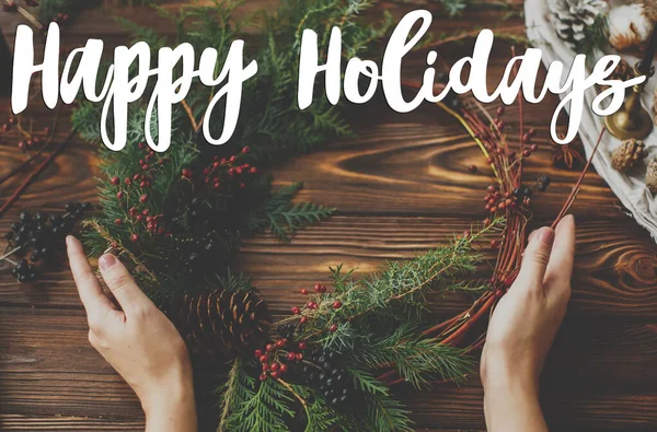 赤い果実と松のコーンと木製のテーブルの上に近代的な素朴なクリスマスの花輪を保持手に手書きの幸せな休日のテキスト グリーティングカード現代の季節の挨拶 — ストック写真