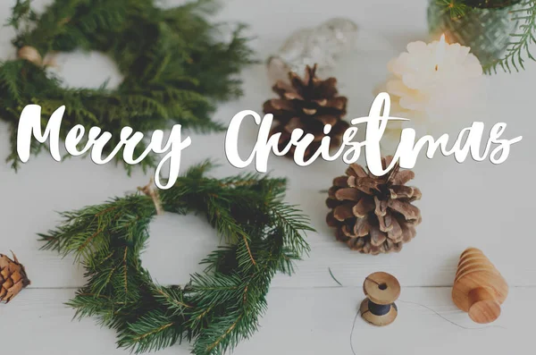 Kırsal Noel çelenginde mumlarla, kozalaklarla ve beyaz ahşap süslemelerle el yazısıyla mutlu Noeller mesajı. Tebrik kartı, modern mevsimler selamları. İyi tatiller.