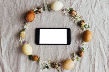 Paskalya çelengi düzlüklerinde boş ekranlı telefon. Paskalya yumurtaları ve bahar çiçekleri rengarenk keten arka planda akıllı telefonlu çelenk, mesaj için yer var. Mevsimler selamlama şablonu