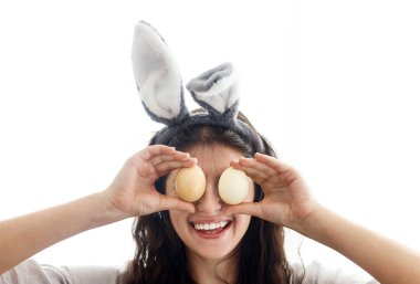 Mutlu Paskalyalar! Tavşan kulaklı ve keten elbiseli mutlu genç kadın paskalya yumurtalarını gözlerinde tutuyor ve beyaz ışıkta gülümsüyor. Doğal boyanmış paskalya yumurtaları, komik anlar.