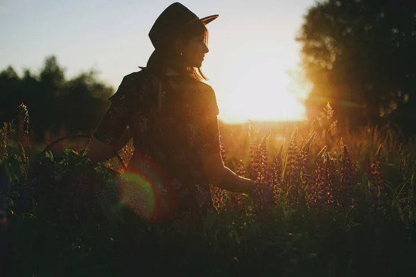 田舎の夕日の光の中でラインを集めるスタイリッシュな女性のシルエット 大気中の静かな瞬間 若い女性が野の花を集め 夏の牧草地でリラックス — ストック写真