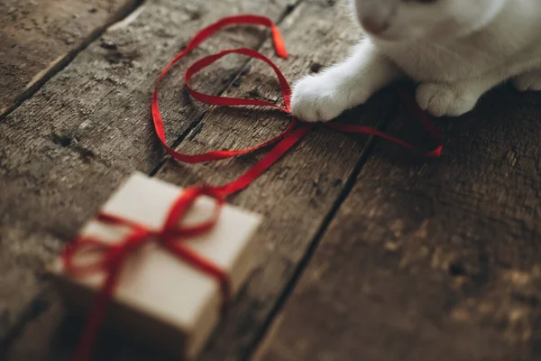 素朴な木製の背景にスタイリッシュなクラフトギフトボックスからの赤いリボンで遊ぶかわいい猫 雰囲気の良いイメージ 絹のリボンにキティの足 ハッピーバレンタインデーと愛の概念 — ストック写真