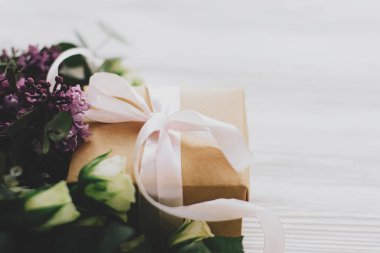Beyaz ahşap arka planda eflatun ve gül buketli şık bir hediye kutusu yumuşak ışıkta, yazı için yer var. Anneler günün ve kadınlar günü konseptin kutlu olsun. Kurdele ve bahar çiçekleriyle birlikte el işi.
