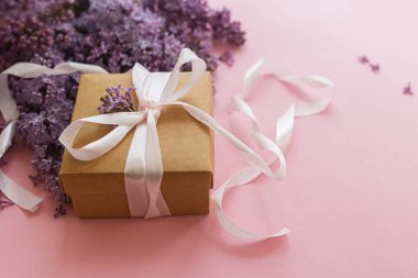 Pembe kağıt üzerinde leylak çiçekleri ve hediye kutusu. Anneler günün ve kadınlar günü konseptin kutlu olsun. Bahar çiçekli tebrik kartı. Mor leylak buketi, şık bir zanaat ve kurdele mevcut.