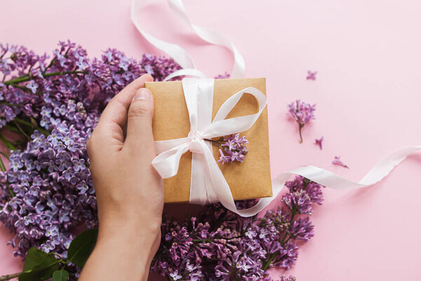 Happy mothers day and valentine's day concept. Hand holding gift box with ribbon and lilac flowers on pink paper, top view. Purple lilac flowers bouquet with craft present box. Giving gift