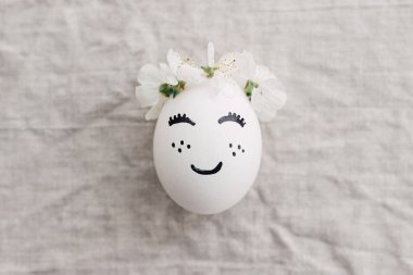 Mutlu Paskalyalar! Doğal yumurta, gülen surat çizilmiş, keten kumaşın üzerinde çiçek tacıyla. Çevre dostu sıfır atık tatil konsepti. Metin için boşluk olan üst görünüm