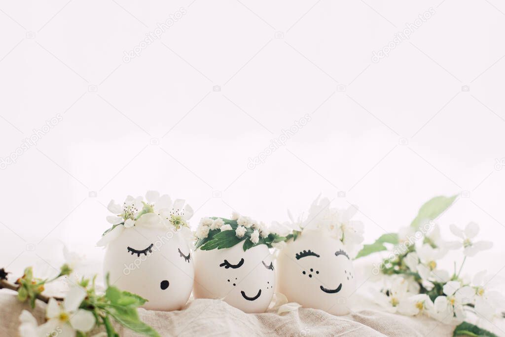 Happy Easter! Natural eggs with drawn cute faces in floral wreaths on linen fabric with blooming spring branch,petals and green leaves in soft light. Space for text. Eco friendly holiday concept