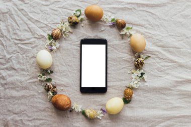Paskalya kırsalı çelengi ve boş ekranlı telefon, metin için boş alan. Paskalya yumurtaları ve bahar çiçekli çelenk. Kırsal keten arka planda akıllı telefon. Mevsimler selamlama şablonu