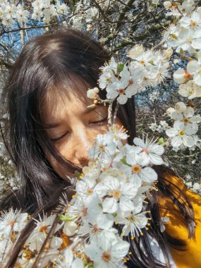 Çiçek açan kiraz dalları arasında poz veren zarif, güzel bir kadın. Şehvetli, sakin bir an. Beyaz çiçeklerle sarılmış sarı ceketli, şık, genç, esmer bir kadın.